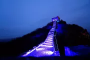 The Great Wall of China illuminated with blue lights, creating a striking visual effect as it curves up a hill toward a watchtower under the evening sky. The surrounding landscape is dark, highlighting the blue-lit pathway that accentuates the ancient stone structure against the quiet backdrop.