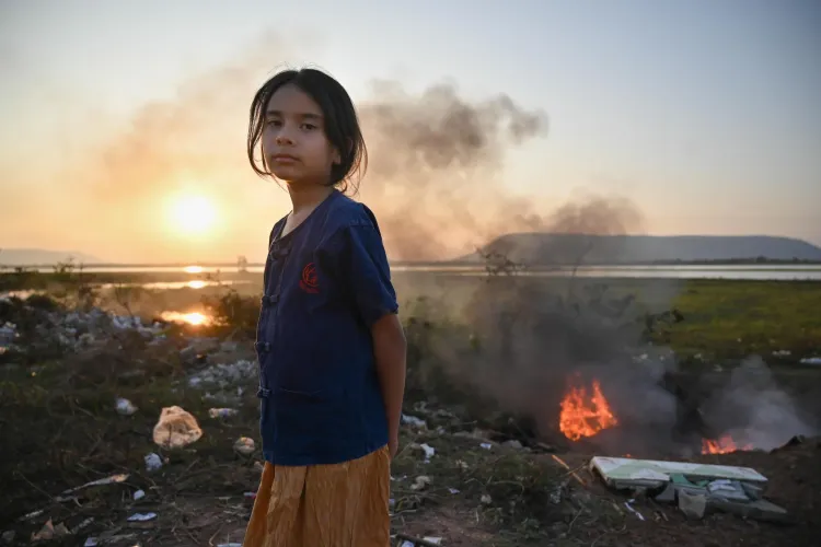 A young girl stands beside a smoky open fire at sunset near a waterside area, with scattered trash and low hills in the background.