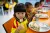 A young girl in a yellow and red shirt sits at a table eating a piece of fruit on a stick, with a metal tray of food in front of her and other children eating in the background.