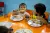 Two young children sit at an orange table eating from metal trays, with one child holding a skewer of banana slices while the other looks on.