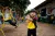 A young girl in a yellow school uniform stands in a playground with her arms crossed, while other children play on climbing equipment and adults sit near a school building behind her.