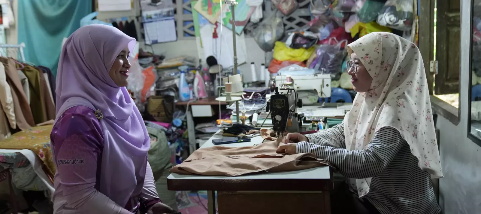 Two women wearing hijabs sit facing each other and smile in a small sewing workshop. One woman is seated at a sewing machine with fabric in front of her, while the other listens attentively. The room is filled with sewing materials, colorful bags, and garments, creating a cozy and busy atmosphere.