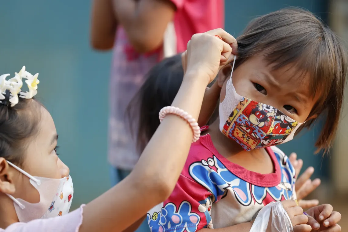 A girl helps another child adjust the ear loop of her colorful face mask while they stand together outdoors.