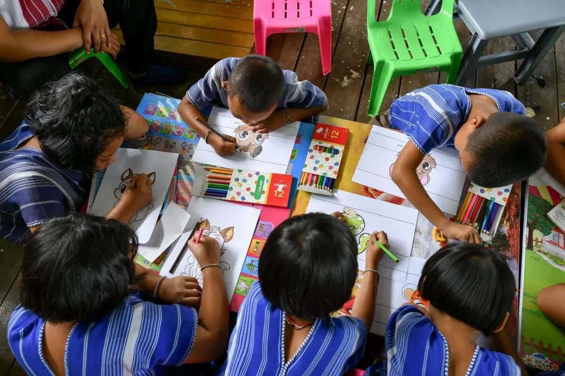 A group of young children sit closely together around a low table, concentrating as they color and draw pictures with crayons during a creative classroom activity.