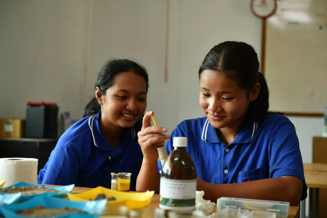 Two girls wearing blue polo shirts are engaged in a science activity in a laboratory. One is holding a test tube while the other smiles beside her. There are various laboratory equipment items on the table.