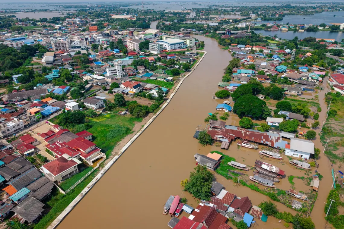 ภาพมุมสูงของเมืองที่น้ำท่วมริมฝั่งแม่น้ำสีน้ำตาล โดยมีบ้านและเรือบางส่วนจมอยู่ในน้ำ
