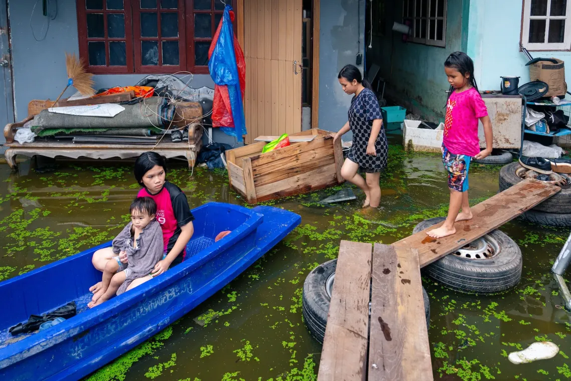 เด็ก ๆ อยู่ในลานบ้านที่ถูกน้ำท่วม โดยมี 2 คนกำลังนั่งเรือสีน้ำเงินและอีก 2 คนเดินบนแผ่นไม้ที่ทำเป็นทางเดินชั่วคราวเหนือระดับน้ำ