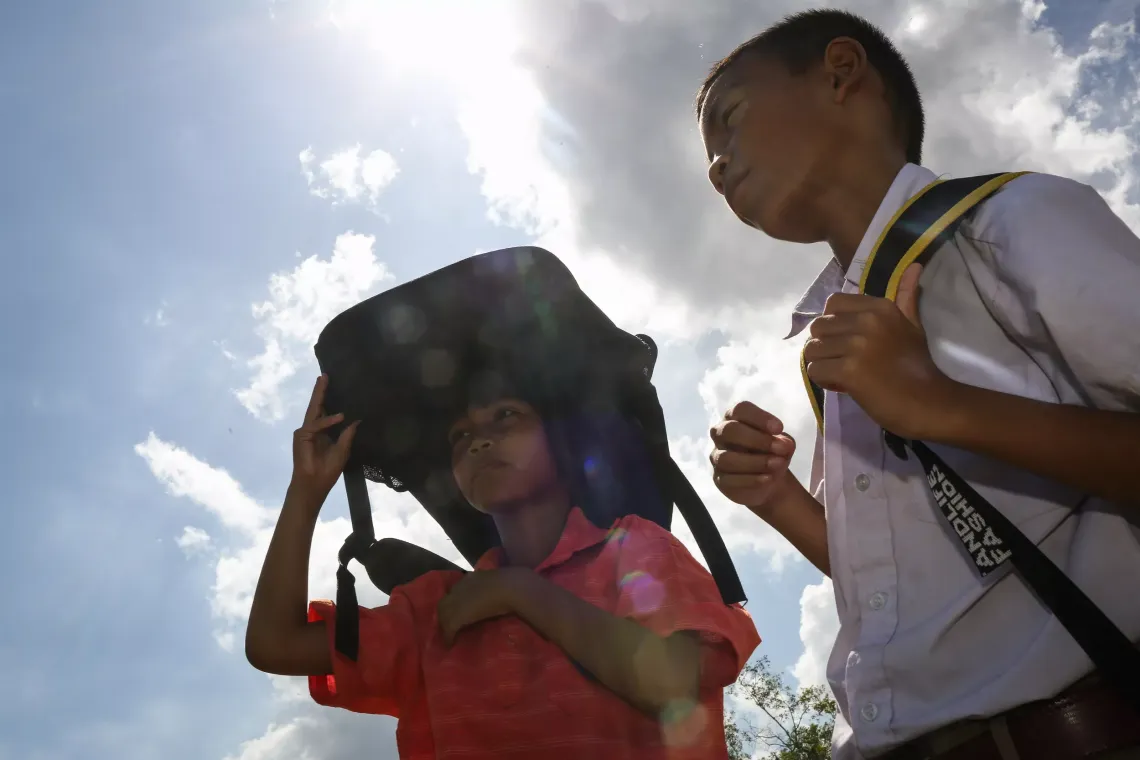 Boy and girl are standing under the sun with school bag cover their heads.