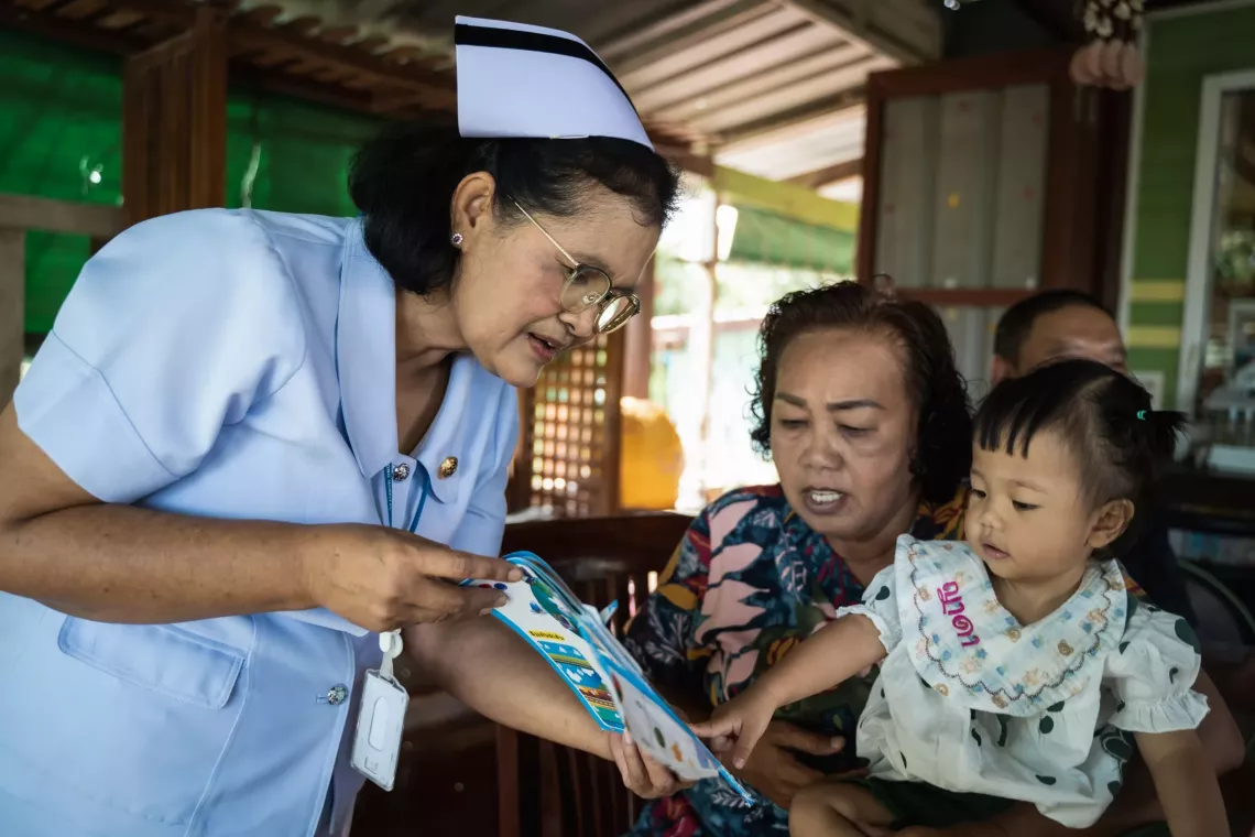 พยาบาลในชุดยูนิฟอร์มสีขาวกำลังแสดงหนังสือภาพให้เด็กหญิงตัวเล็กและผู้ดูแลดูระหว่างการเยี่ยมบ้าน เด็กนั่งอยู่บนตักของผู้ดูแลและชี้ไปที่หนังสือด้วยความสนใจ ขณะที่พยาบาลโน้มตัวเข้ามาอย่างตั้งใจ