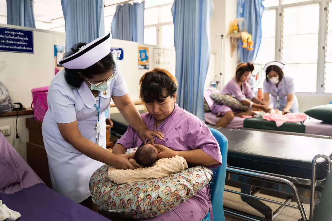 พยาบาลสวมชุดขาวและหน้ากากอนามัยกำลังช่วยคุณแม่มือใหม่ให้นมบุตรในหอผู้ป่วยของโรงพยาบาล คุณแม่นั่งอยู่บนเก้าอี้ สวมชุดคนไข้สีม่วง และอุ้มลูกน้อยโดยมีหมอนหลากสีรองรับอยู่ ด้านหลังมีพยาบาลอีกคนหนึ่งกำลังดูแลคุณแม่และทารกอีกคนบนเตียงในห้องที่มีม่านสีน้ำเงินและแสงธรรมชาติจากหน้าต่างบานใหญ่