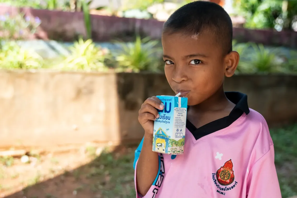 A young boy in a pink school uniform drinks from a blue carton of milk with Thai text, standing outdoors near a garden wall.