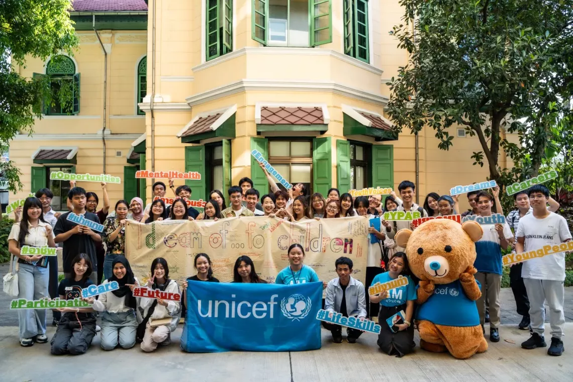 A group of young people and UNICEF staff pose in front of a yellow building with green windows, holding signs with positive hashtags like "#Opportunities," "#MentalHealth," "#YouthVoices," and "#Dreams." They display a banner reading "I dream of...for children," signed by participants. A UNICEF Teddy Blu mascot stands with them, creating a cheerful and hopeful atmosphere.