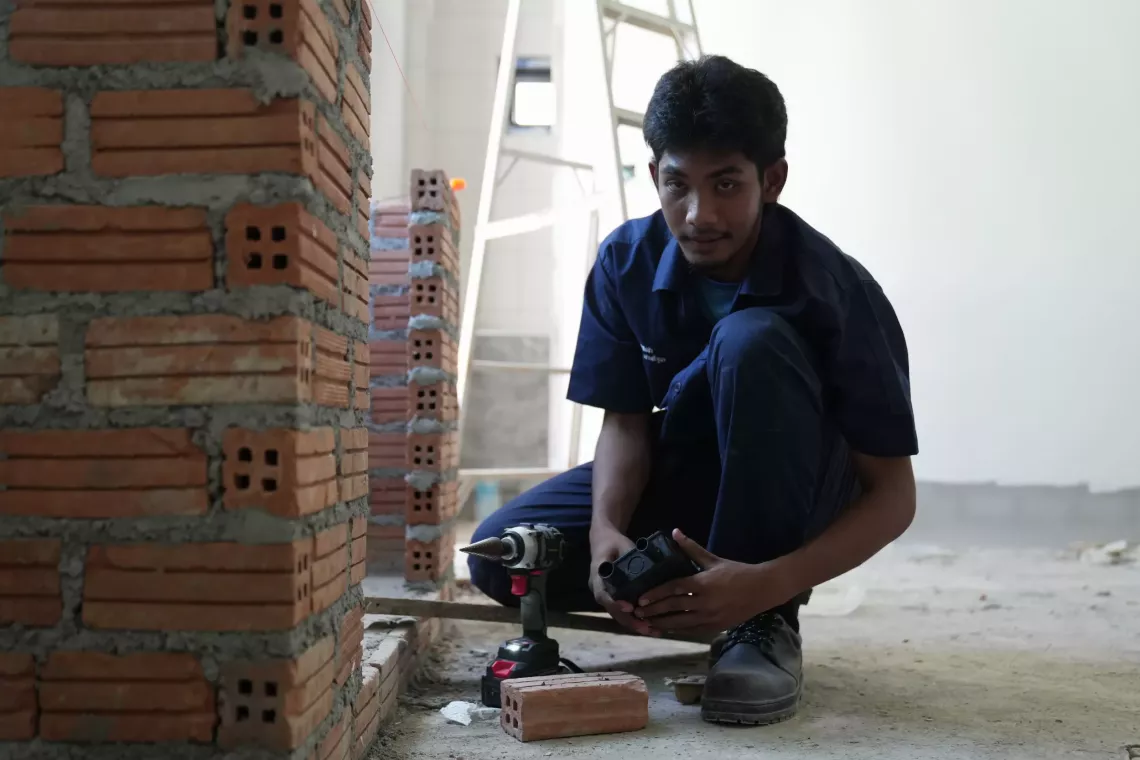 A young intern in a dark uniform crouches beside a brick wall under construction, holding tools with a power drill on the floor nearby.