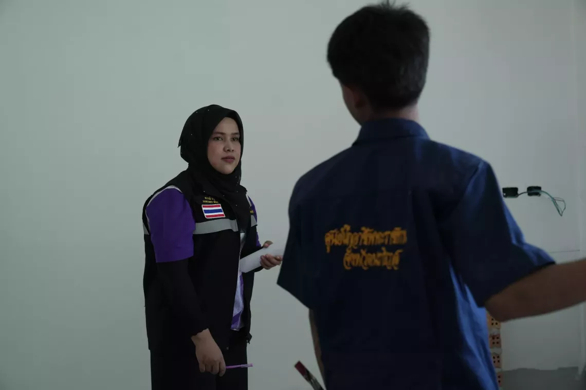 A woman in a hijab and uniform holds a pen and paper while talking to a young intern in a blue vocational training uniform inside a construction area.