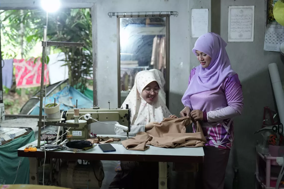 Two women in hijabs smile while working together on a brown garment at a sewing table in a modest indoor workshop.