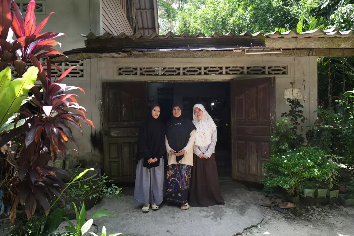 Three women wearing hijabs stand together in front of an old wooden house surrounded by lush green plants, smiling in natural sunlight.