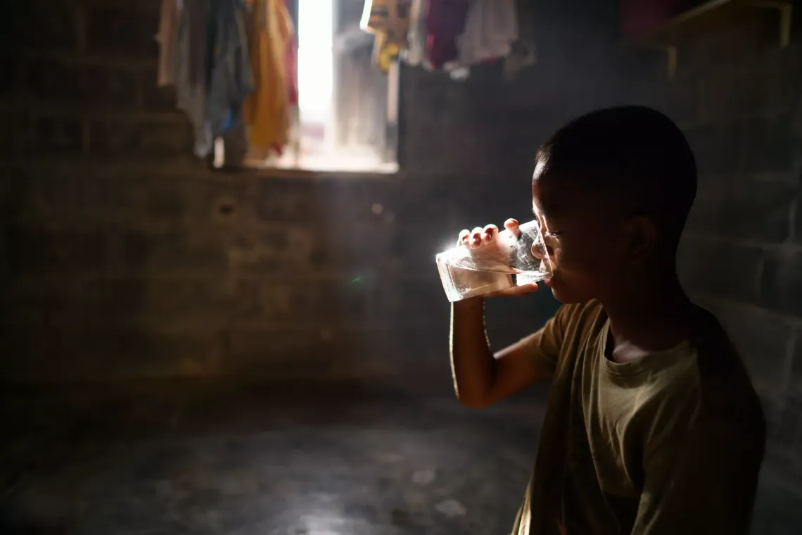 A young boy drinks water from a glass in a dimly lit room with concrete walls. Sunlight streams in through a small window in the background.