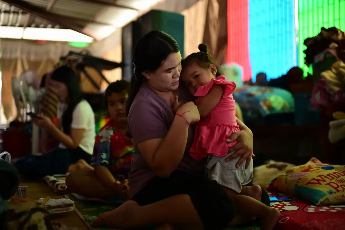 A mother sits on a mat holding her daughter close to her chest inside a crowded shelter, with other families resting in the background.