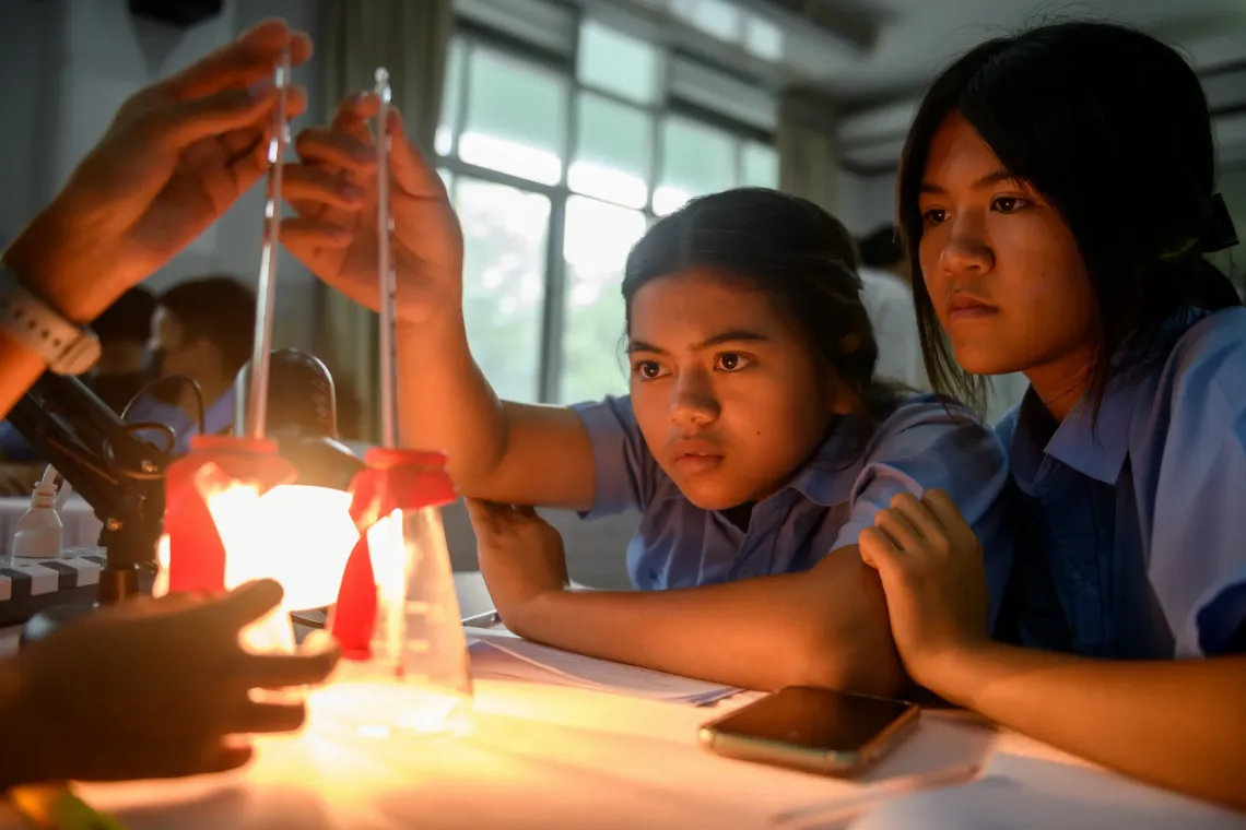 Two schoolgirls work together during a science lesson, carefully observing a simple experiment with measuring tubes and glassware under a teacher&rsquo;s guidance in a classroom.