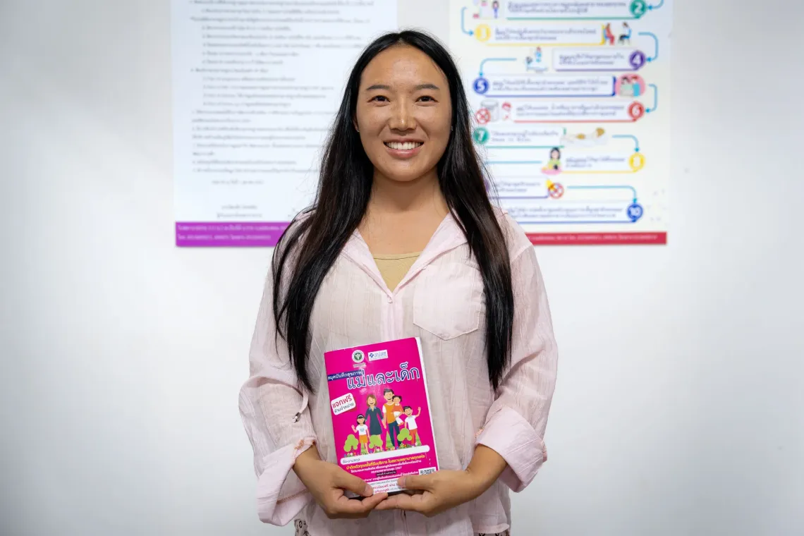 Daraporn Yoopao, smiling and holding a pink booklet titled in Thai &ldquo;monthe and child&rdquo; standing in front of informational posters on a wall.