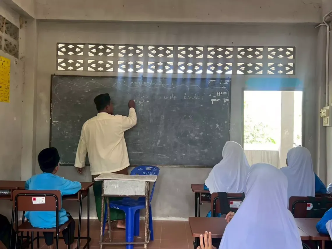 A teacher writing on a chalkboard in a classroom with students in blue uniforms, some of whom are wearing white hijabs.