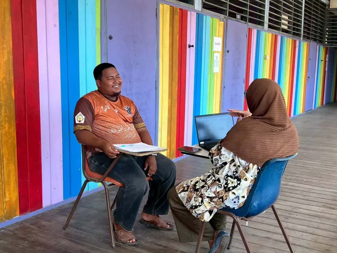 Two people having a conversation in front of a wall painted with colorful vertical stripes; one is holding documents, and the other is using a laptop.