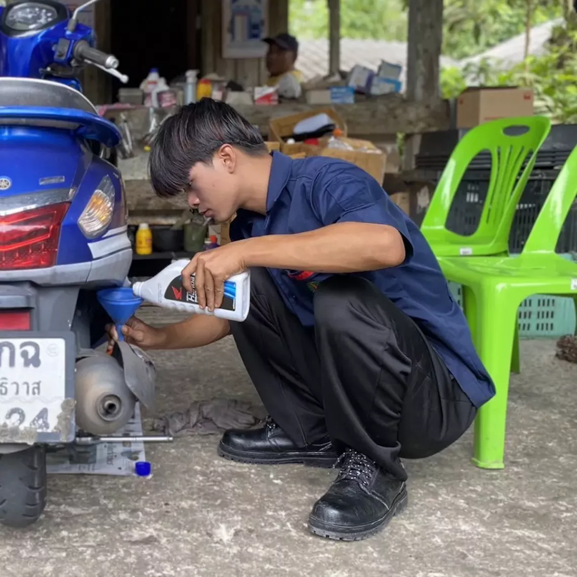 A young intern in a dark uniform crouches beside a blue motorcycle, carefully pouring oil through a funnel during maintenance work.