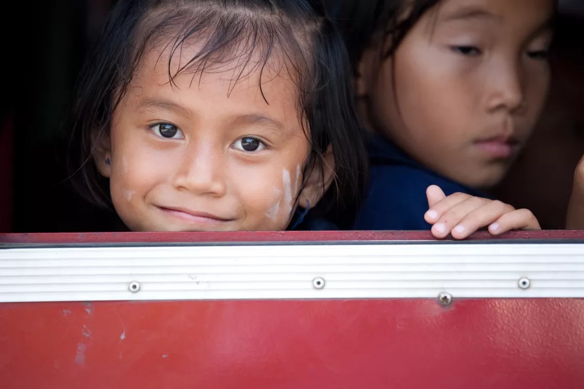 Two young girls are looking out of the window. One of them is looking at the camera, smiling.