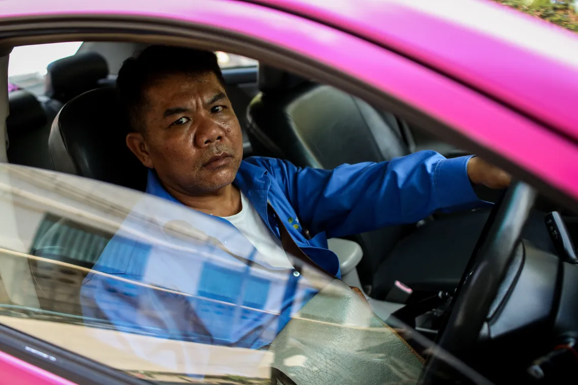 A middle-age taxi driver sitting inside a magenta-colored taxi.
