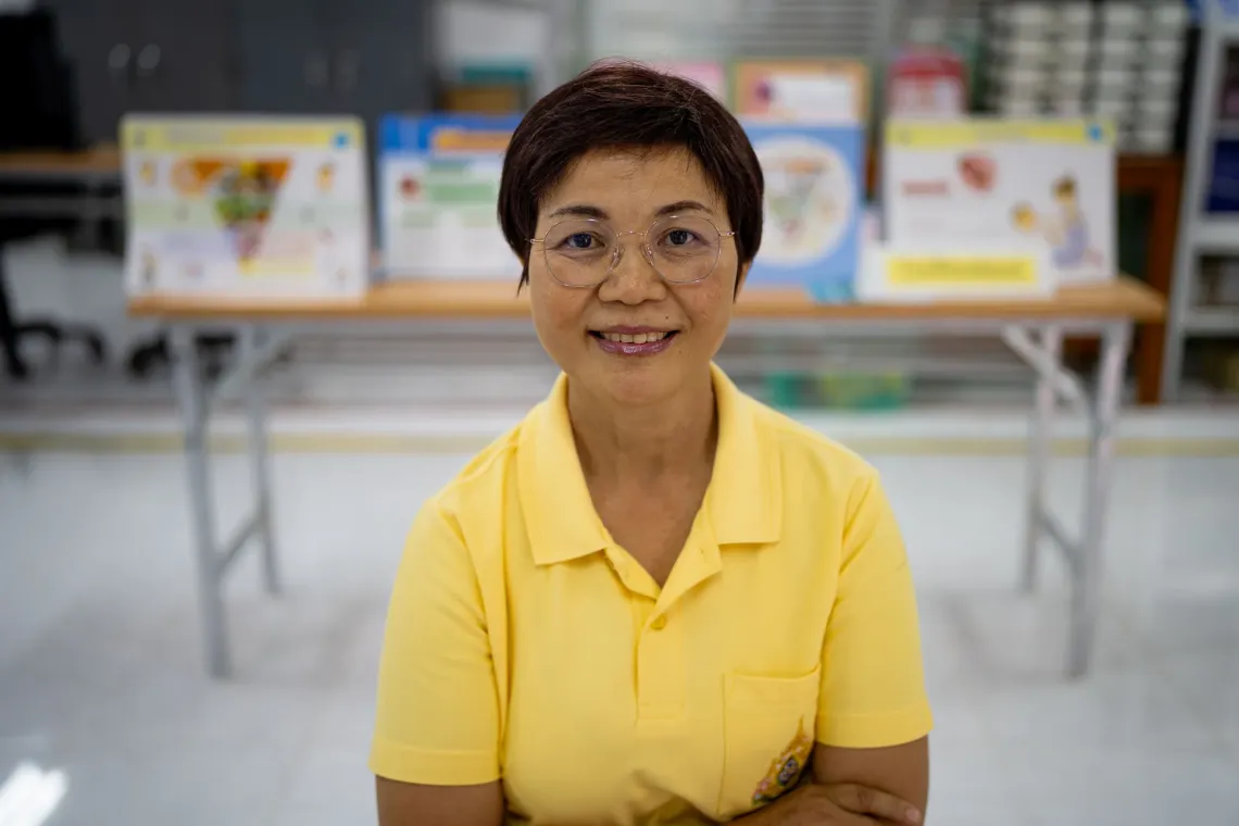 Jampoon Promwangsri, a senior nurse, wearing a yellow polo shirt and glasses smiling in a bright room with educational posters in the background.