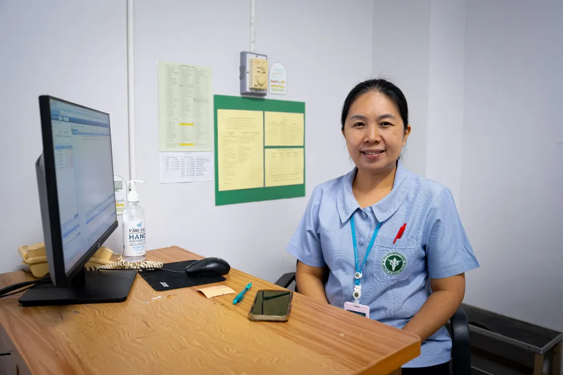 Kanjana Techawaraporn, a nurse, in a light blue uniform sitting at a desk with a computer, sanitizer bottle, and paperwork in a clinical office setting.