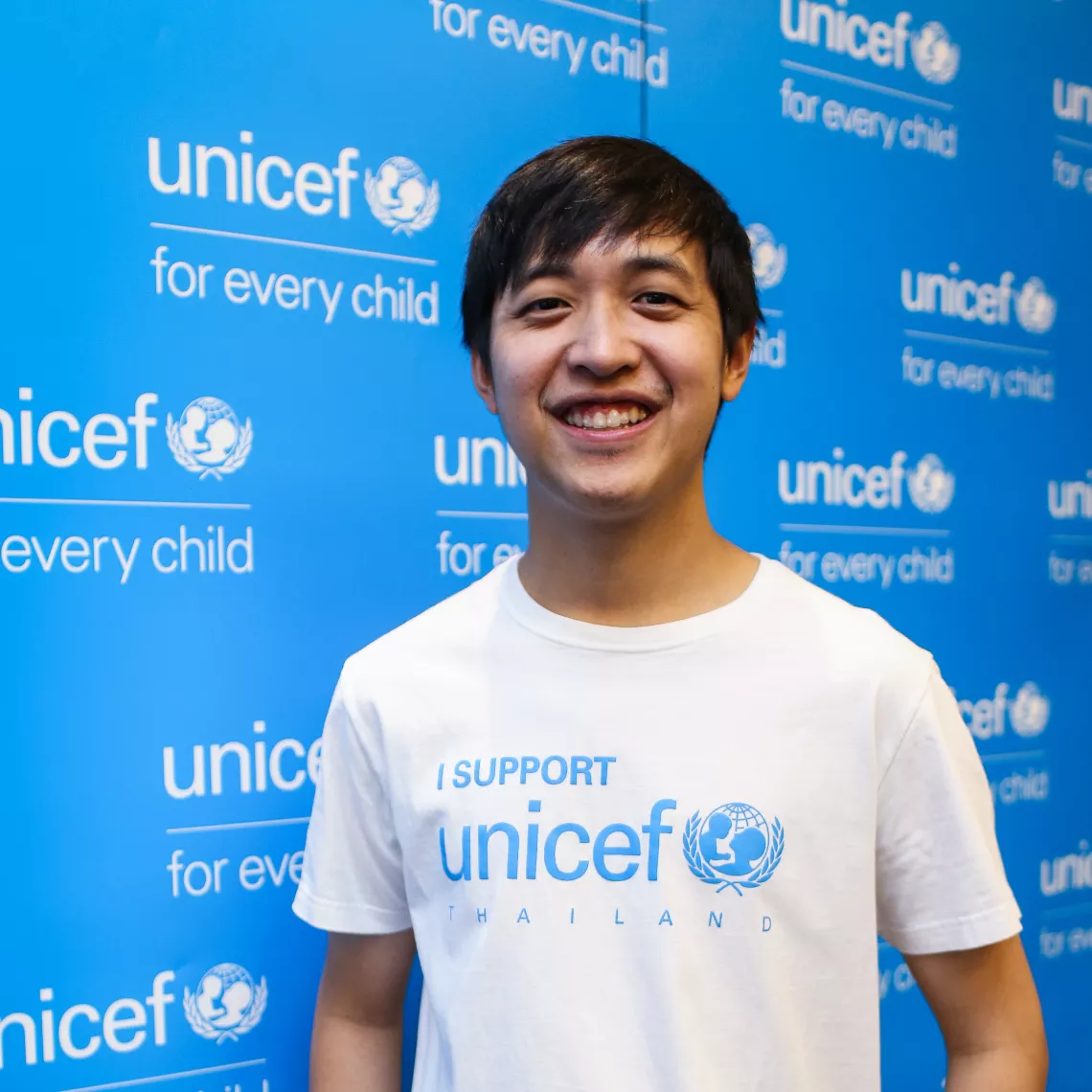 ชายหนุ่มใส่เสื้อยืนสีขาว มีสกรีนเขียนว่า "I support UNICEF Thailand" กำลังยืนอยู่หน้าแบ็กดรอปสีฟ้าของยูนิเซฟ