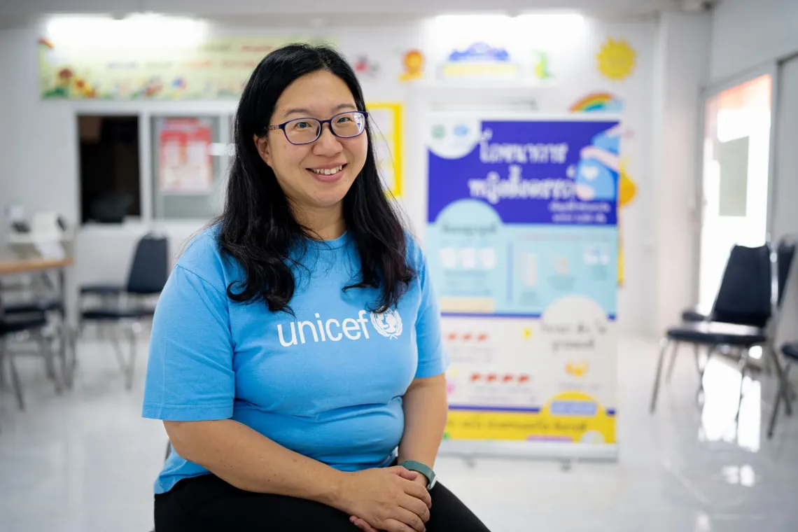 Napat Phisanbut, an Early Childhood Development specialist at UNICEF Thailand, wearing a blue UNICEF T-shirt and glasses sitting in a bright room with informational posters in the background.