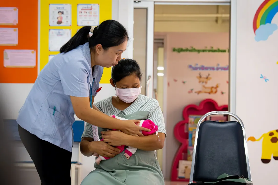 A nurse assisting a masked woman holding a baby doll in a brightly colored clinic room decorated with children's illustrations.