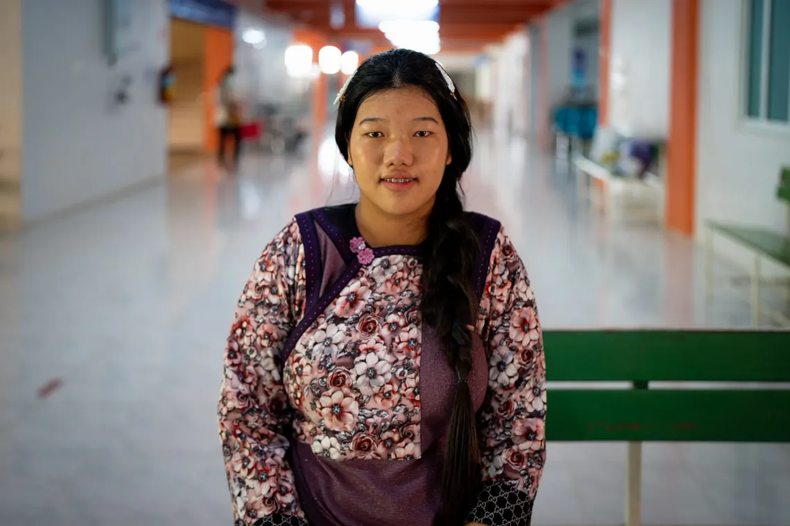 Sirisakorn Laolee, wearing a floral-patterned top with a long braided hairstyle sitting in a bright hallway of a hospital.