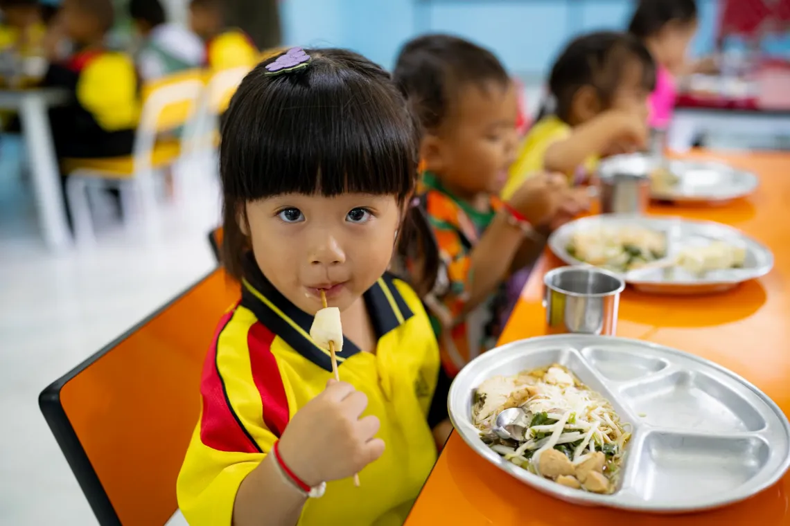 A young girl in a yellow and red shirt sits at a table eating a piece of fruit on a stick, with a metal tray of food in front of her and other children eating in the background.