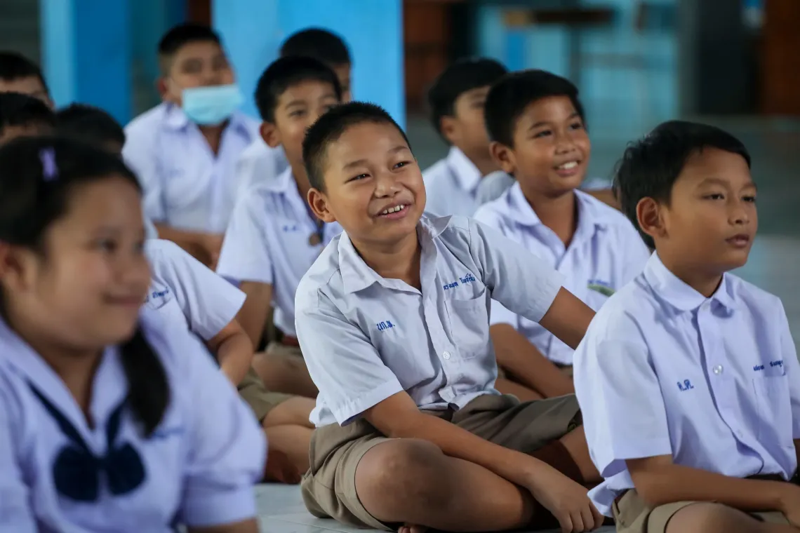 A group of primary school students in white uniforms sit together on the floor, smiling.
