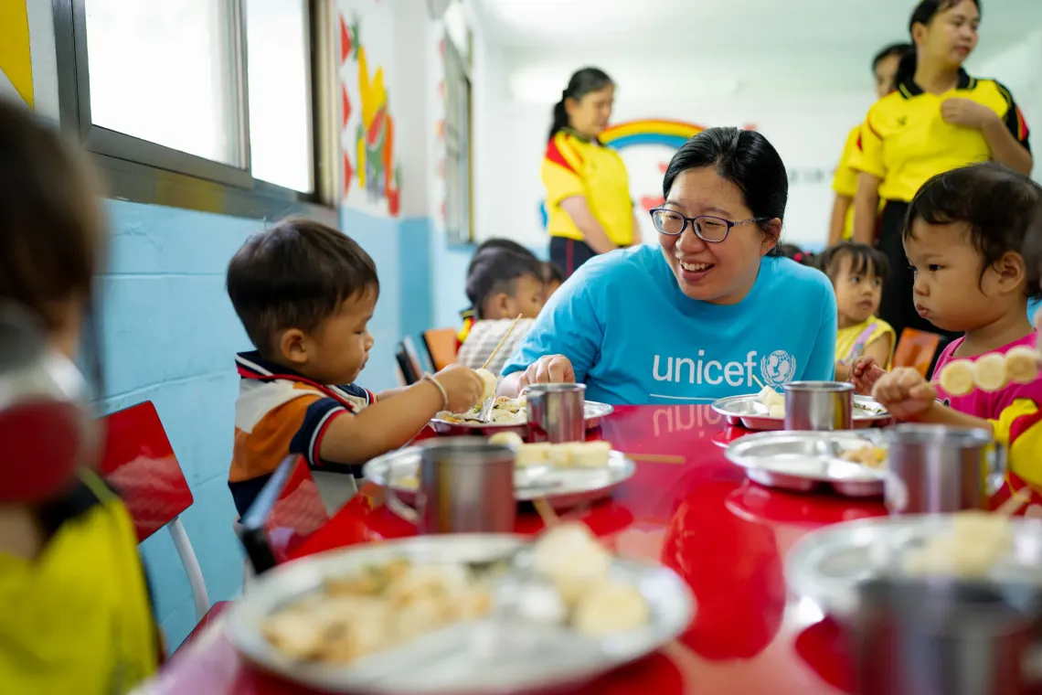 A UNICEF staff member in a blue shirt sits at a red table smiling with young children as they eat from metal trays in a childcare center.