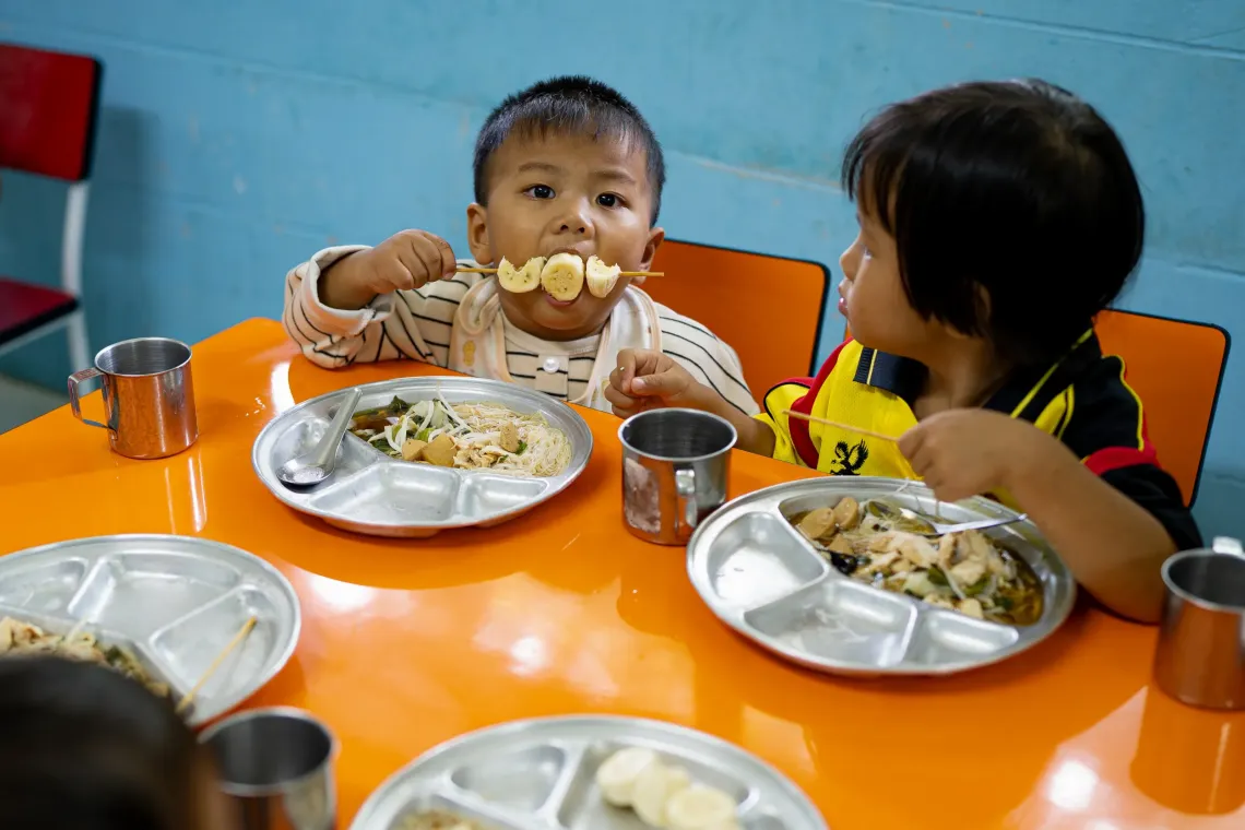 Two young children sit at an orange table eating from metal trays, with one child holding a skewer of banana slices while the other looks on.