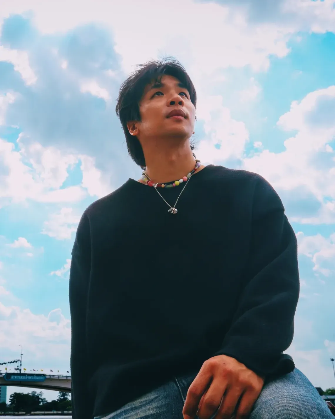 TangBadVoice in a black sweater and layered necklaces looks upward against a bright blue sky filled with clouds.