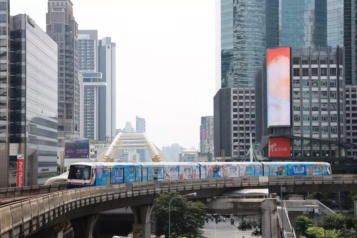 A train on an elevated BTS Skytrain track in Bangkok is wrapped in UNICEF visuals promoting the "A Better Tomorrow Today" campaign. The background features modern skyscrapers, including the distinctive golden arch of the Chong Nonsi Skywalk. Below, traffic moves along the street amidst trees and urban structures.