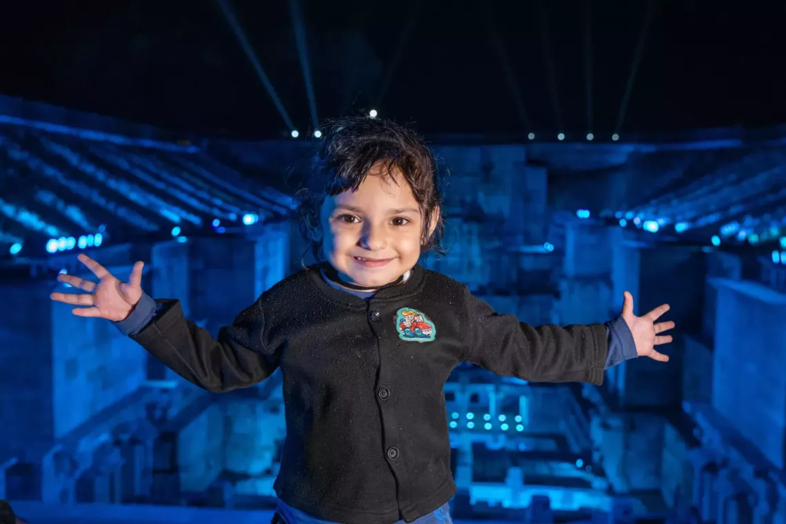 A young child with dark, curly hair smiles widely with arms outstretched, standing against a backdrop of a blue-lit arena or stadium at night. The child wears a black jacket with a colorful patch on the chest, and the scene is illuminated by bright lights, creating a joyful and vibrant atmosphere.