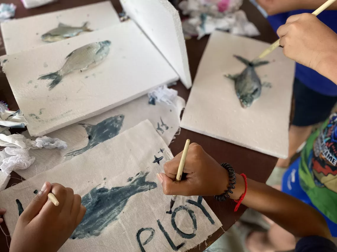 Children are engaged in a fish-printing activity, using real fish coated with paint and pressed onto fabric or paper. Hands holding brushes are seen adding details around the fish prints. The table is scattered with styrofoam boards, painted fish, and cloths.