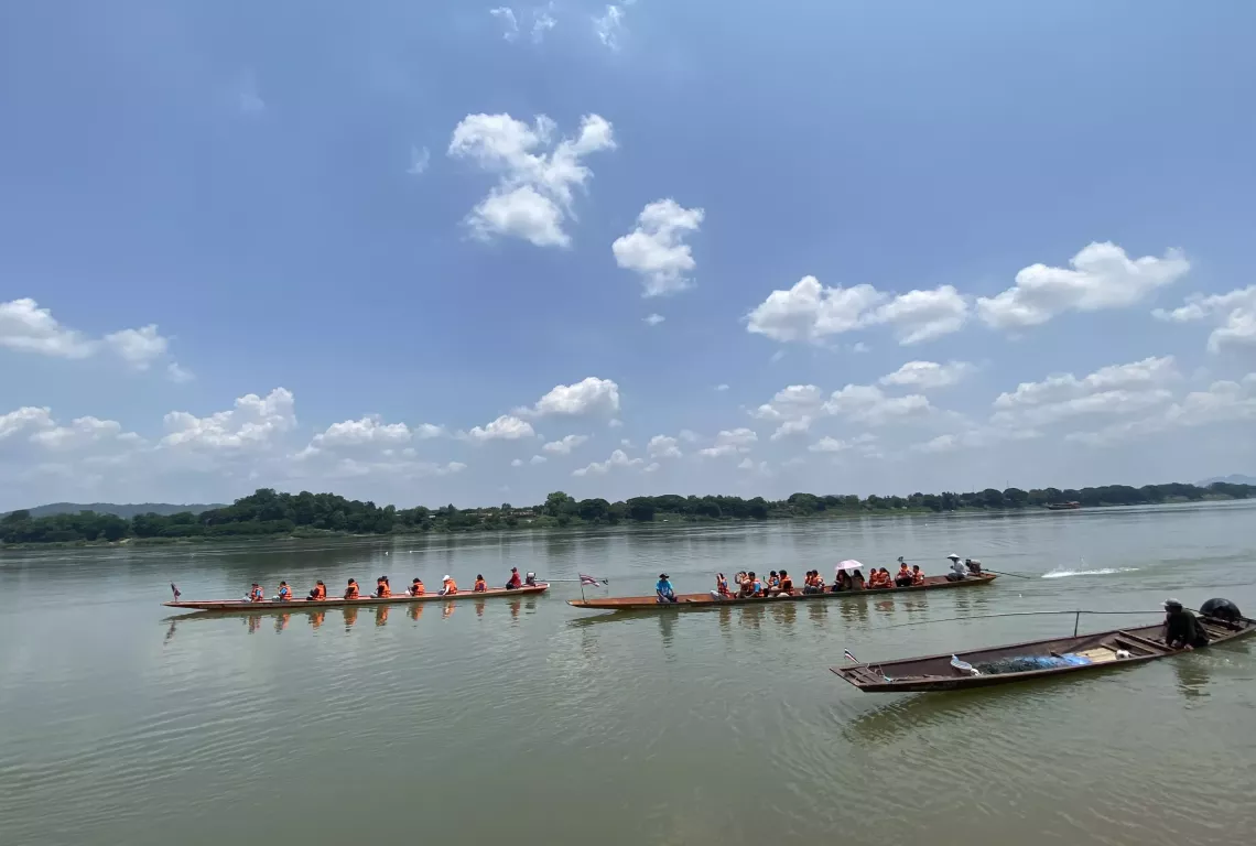 Three long wooden boats are seen floating on a calm river under a bright blue sky. Two boats are filled with people wearing orange life vests, while the third smaller boat in the foreground carries a single person operating it. The background shows a lush green riverside landscape.