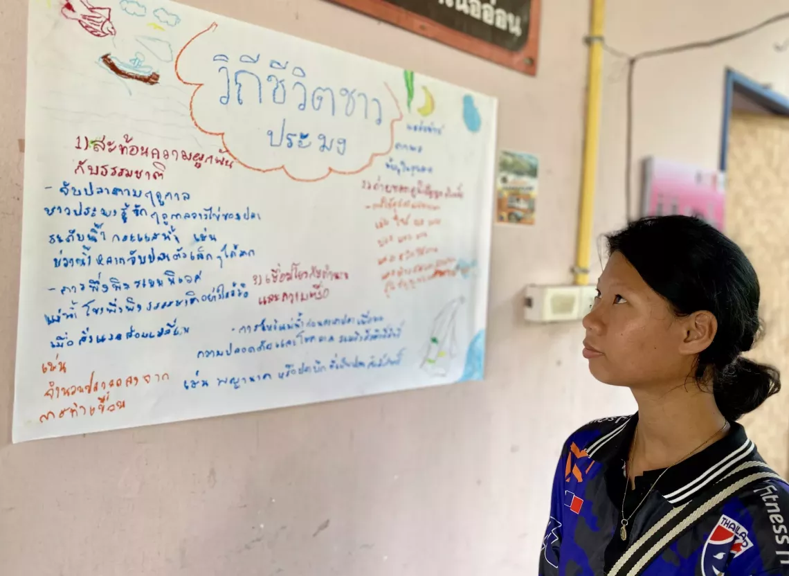 A girl stands indoors and attentively reads a large handwritten poster on the wall titled &ldquo;Fisherfolk&rsquo;s Way of Life". The poster is written in colorful Thai script and includes drawings of a boat, fish, and net, with various points about fishing traditions and practices.