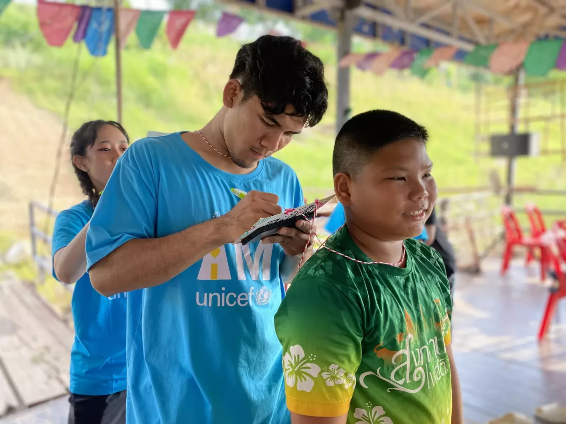 A young man in a blue I Am UNICEF T-shirt writes on a notepad while tying a string necklace with a name tag around a boy&rsquo;s neck. The boy, wearing a green shirt with a floral design, smiles as he participates. Another young woman in blue assists in the background. The activity takes place in a covered open-air space decorated with colorful flags.