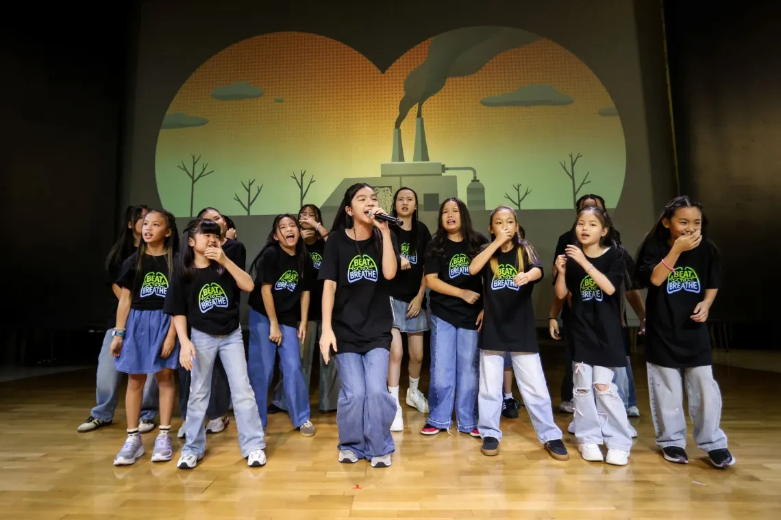 Children wearing &ldquo;Beat to Breathe&rdquo; campaign T-shirts sing together on stage, with an illustration of a smoke-emitting factory and barren trees behind them.