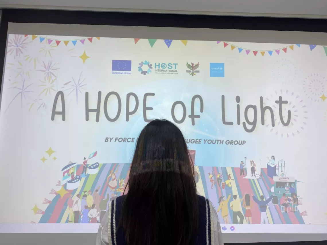A young woman with long dark hair is standing in front of a projector screen displaying a presentation slide titled "A HOPE of Light". The slide features colorful illustrations of people celebrating, along with logos of the European Union, HOST International, and UNICEF. The setting is indoors, likely at an event or workshop.
