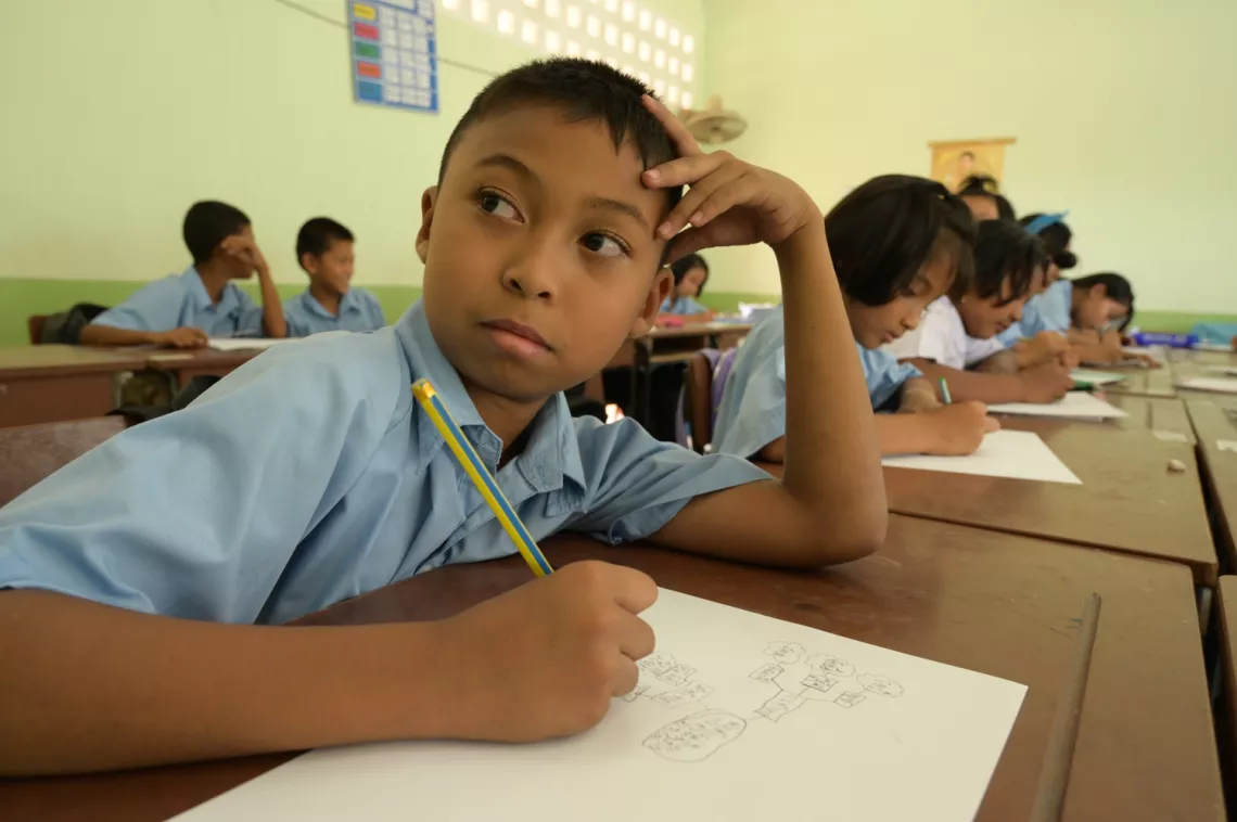 A group of students sitting in a class. One boy is looking at something while taking notes.