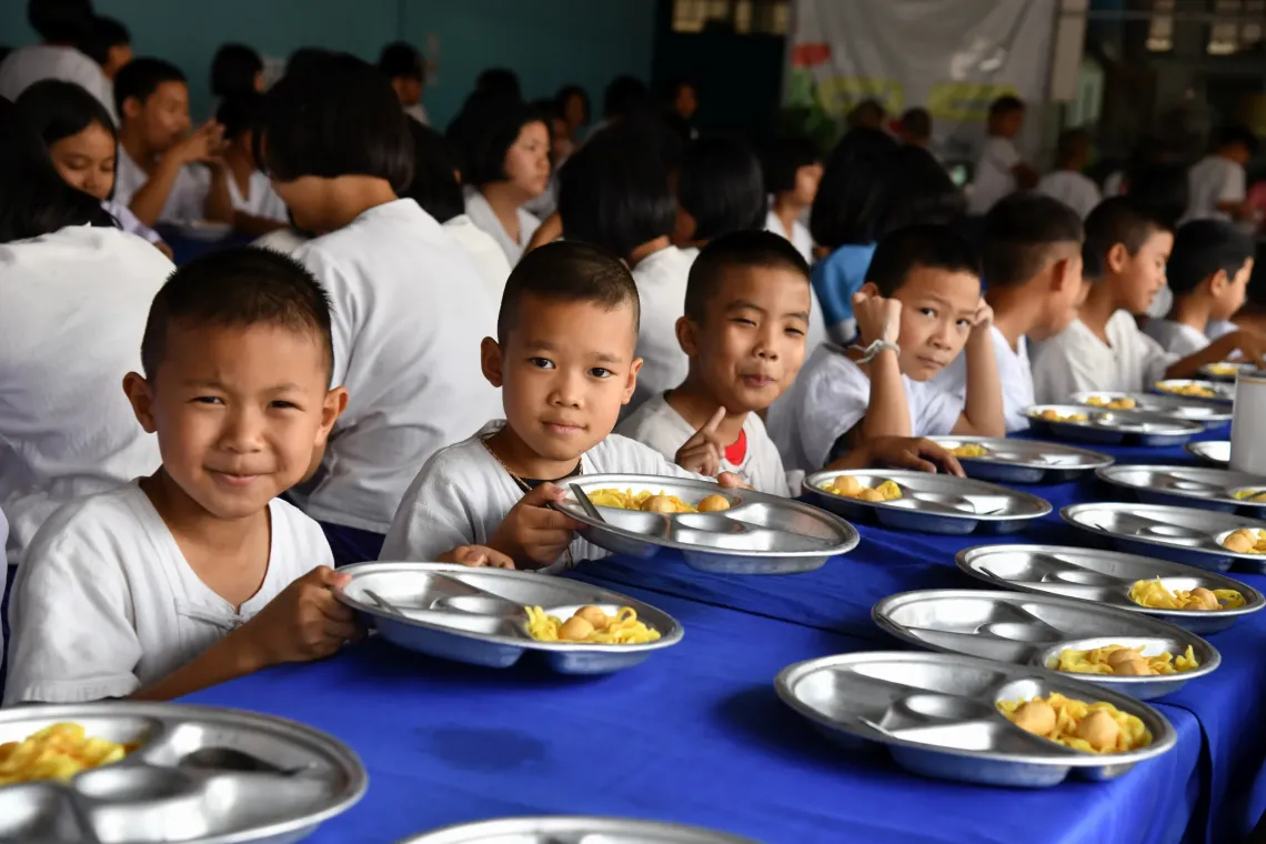 Several young boys sit at a long table with metal lunch trays, looking toward the camera in a crowded school dining hall.